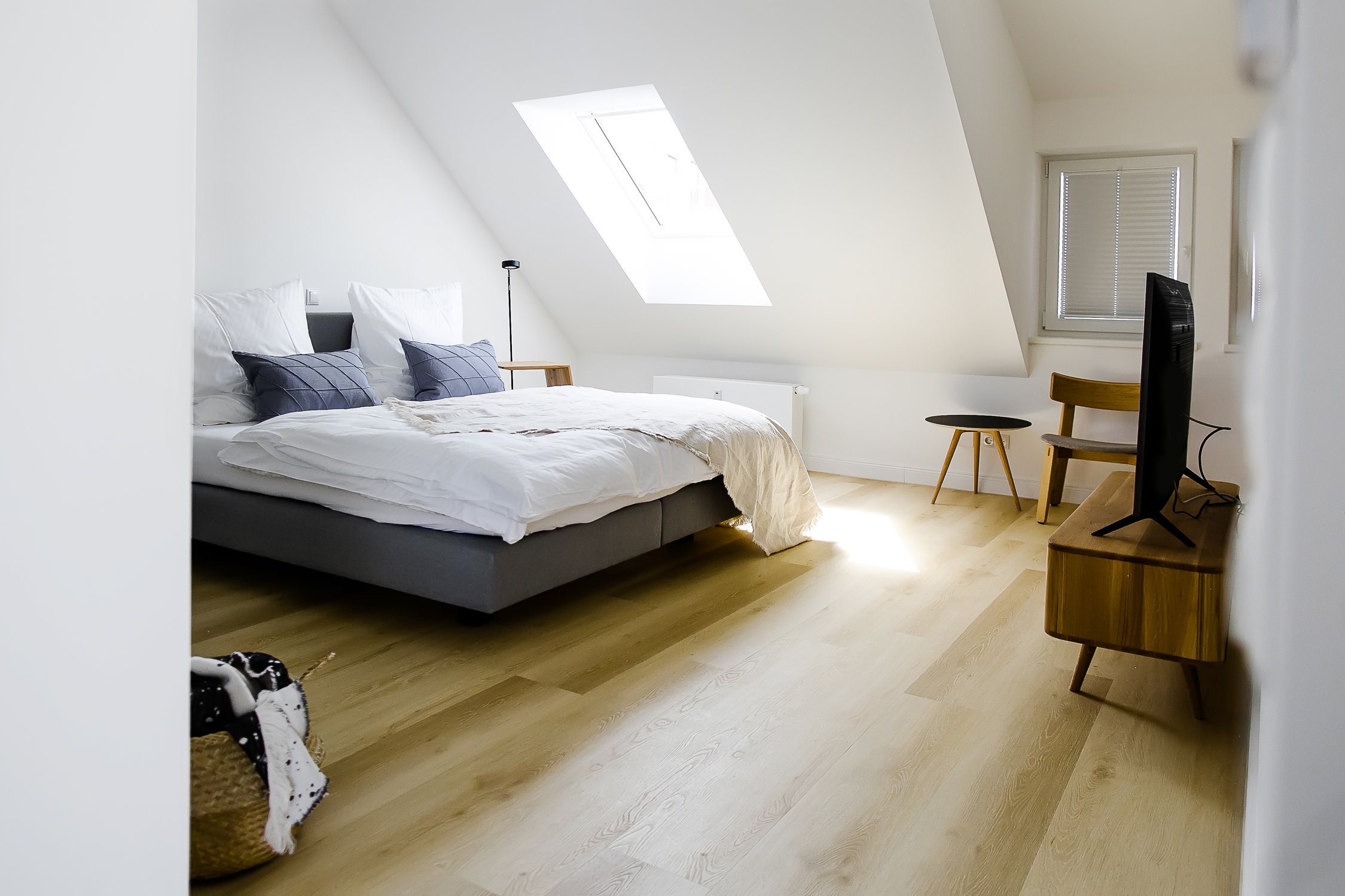 Modernes Dachzimmer mit Dachfenster, Bett mit blauen Kissen, Holzmöbeln im Mid-Century-Stil und minimalistischer Einrichtung auf Holzboden