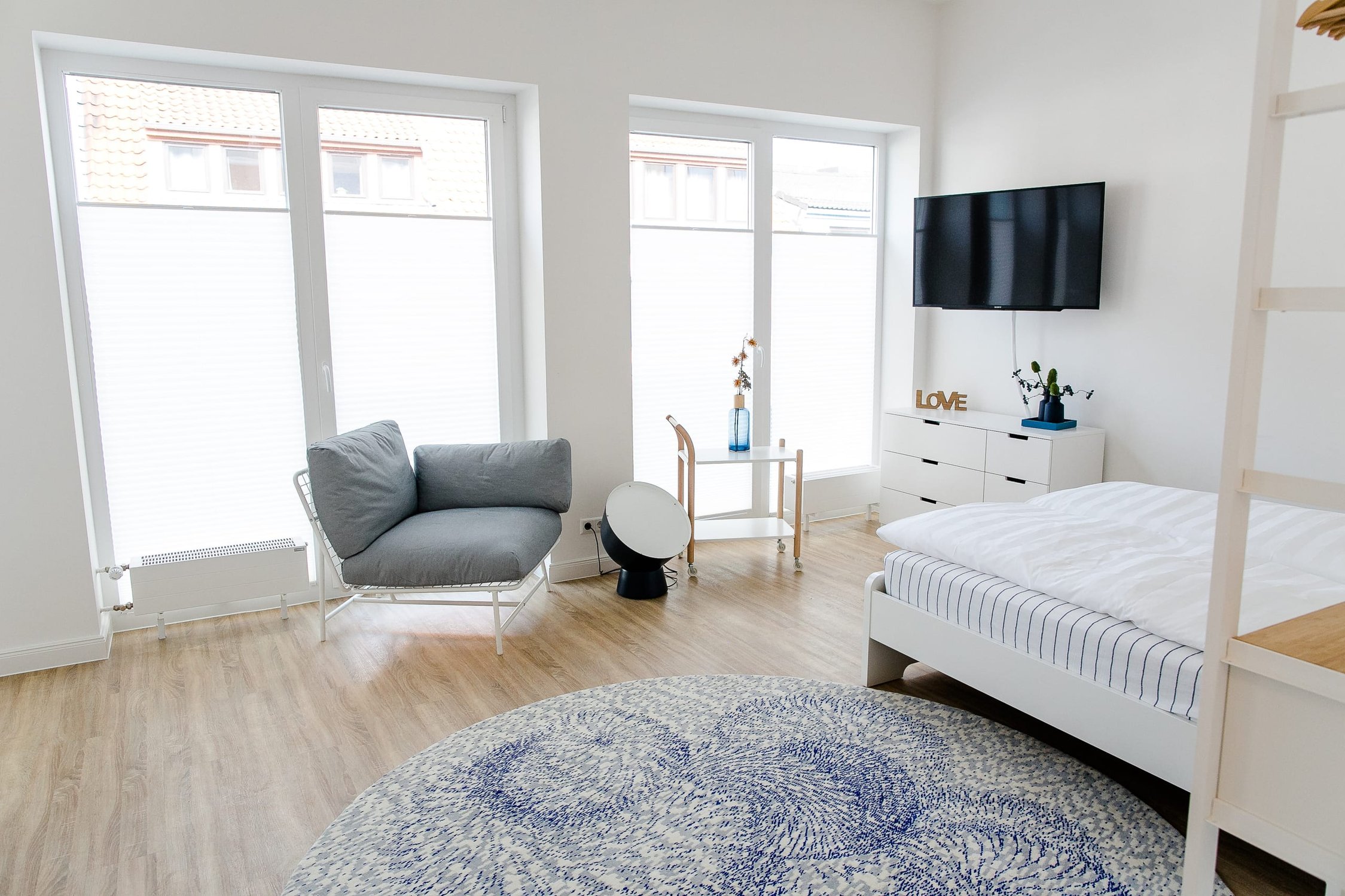 Ein helles Schlafzimmer mit Eichenholzboden, Fenster und weißen Möbeln, einem Sofa und blauem Teppich