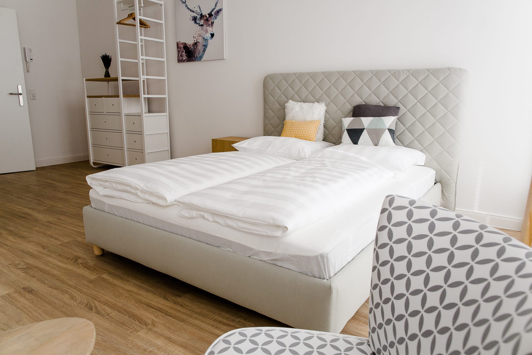 Schlafzimmer mit weißem Bett, gepolstertem Kopfteil, weißem Regal und Holzfußboden