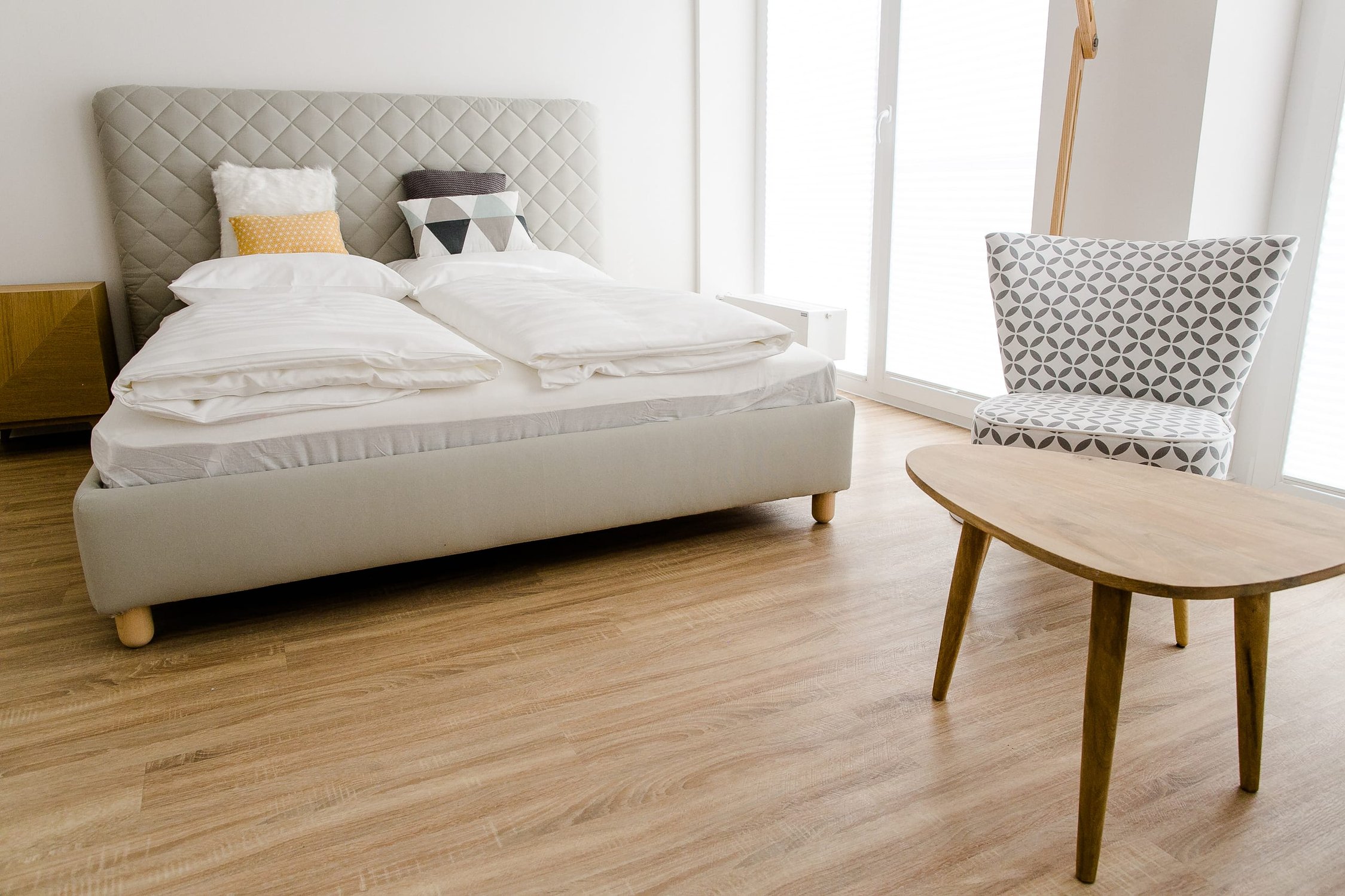 Schlafzimmer mit hellgrauem Bett, weißen Bezügen und Holzboden, heller Raum