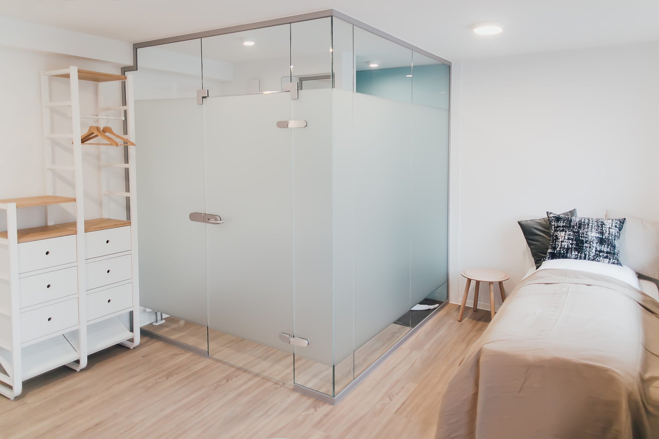 Heller Raum mit milchiger Glasduschkabine, weißem Regal mit Holzeinlagen, Bett und Hocker