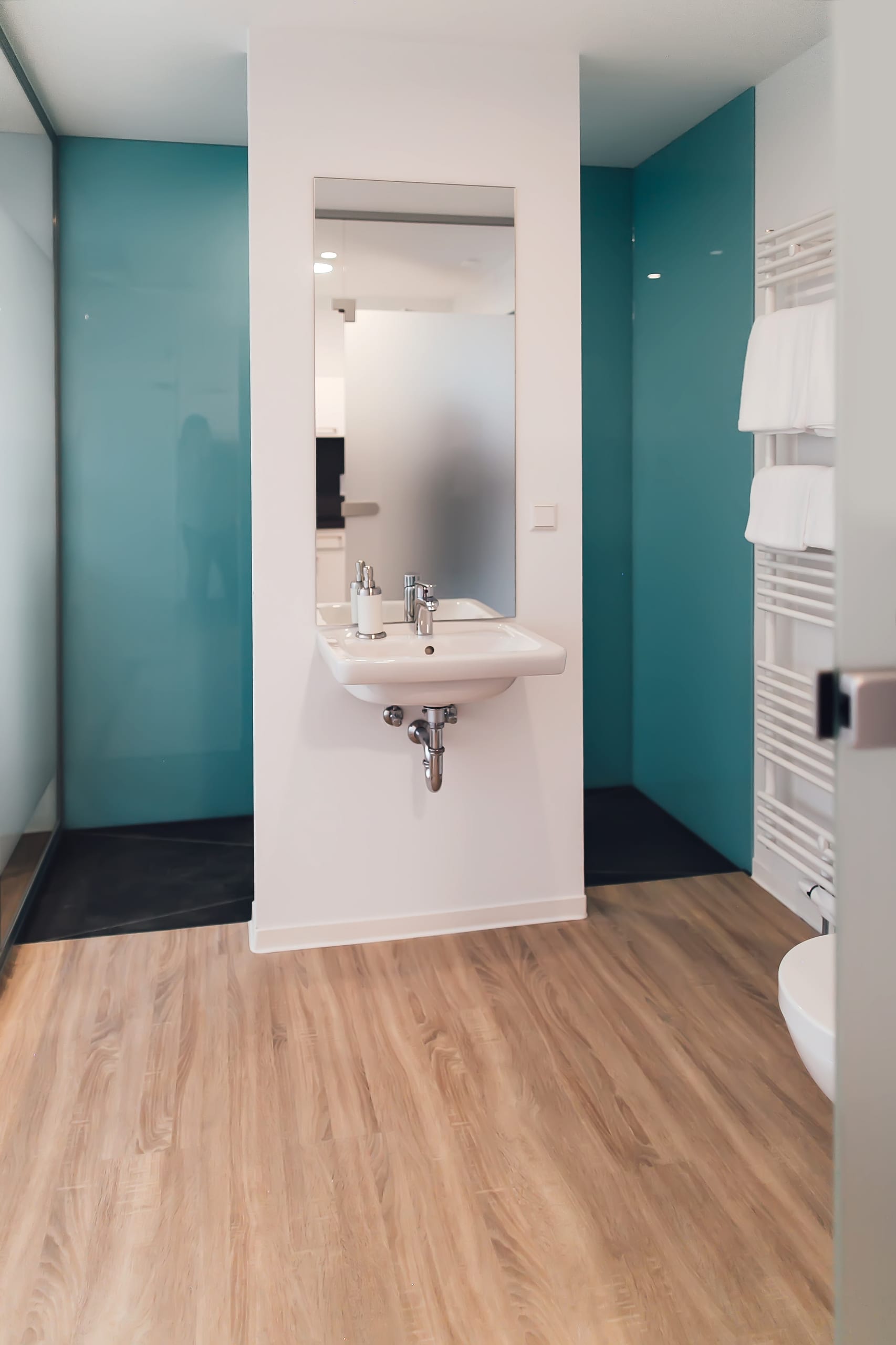 Badezimmer mit Holzoptik-Boden, Spülbecken, Spiegel und türkisen Wänden, weißer Handtuchtrockner