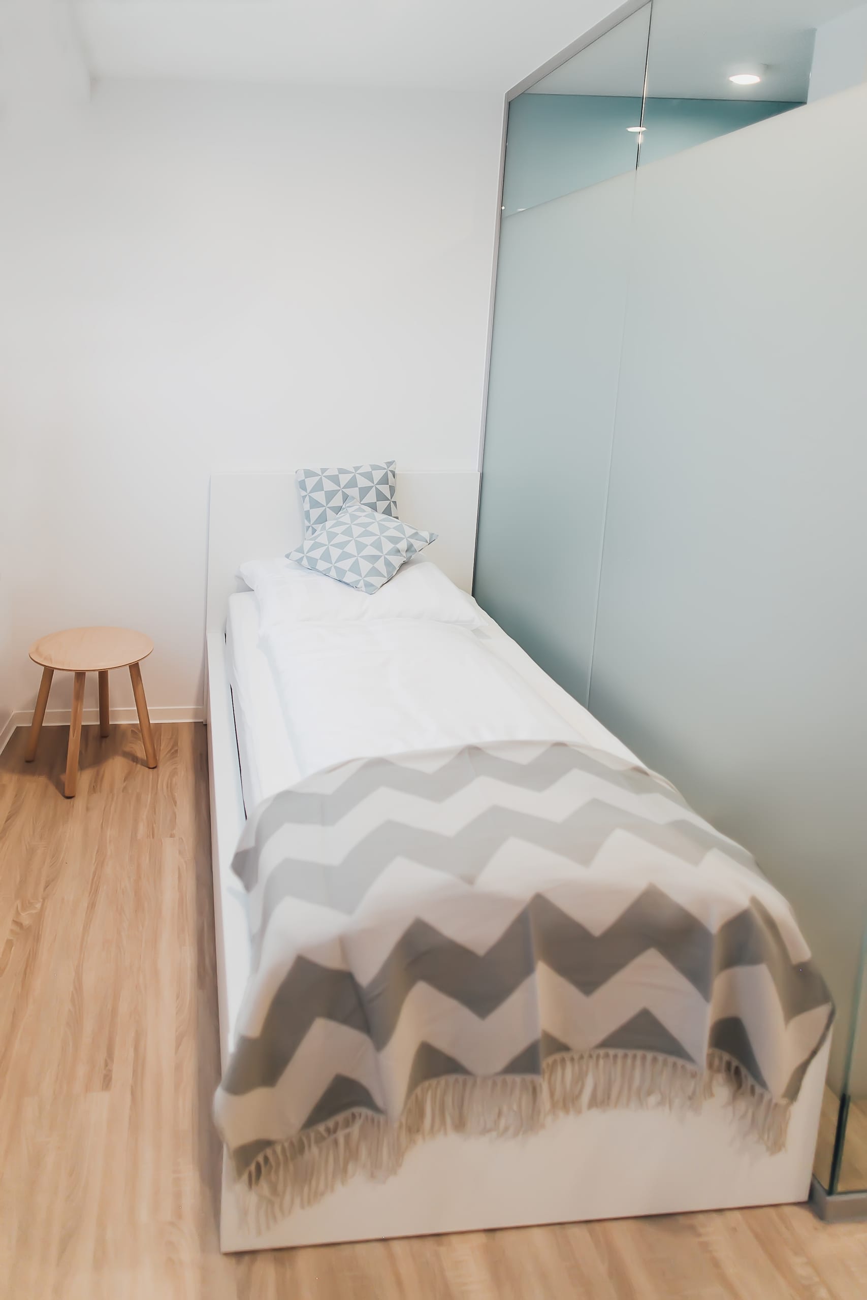 Schlafzimmer mit weißem Einzelbett, grauer Zickzackdecke, hellem Holzfußboden und Hocker