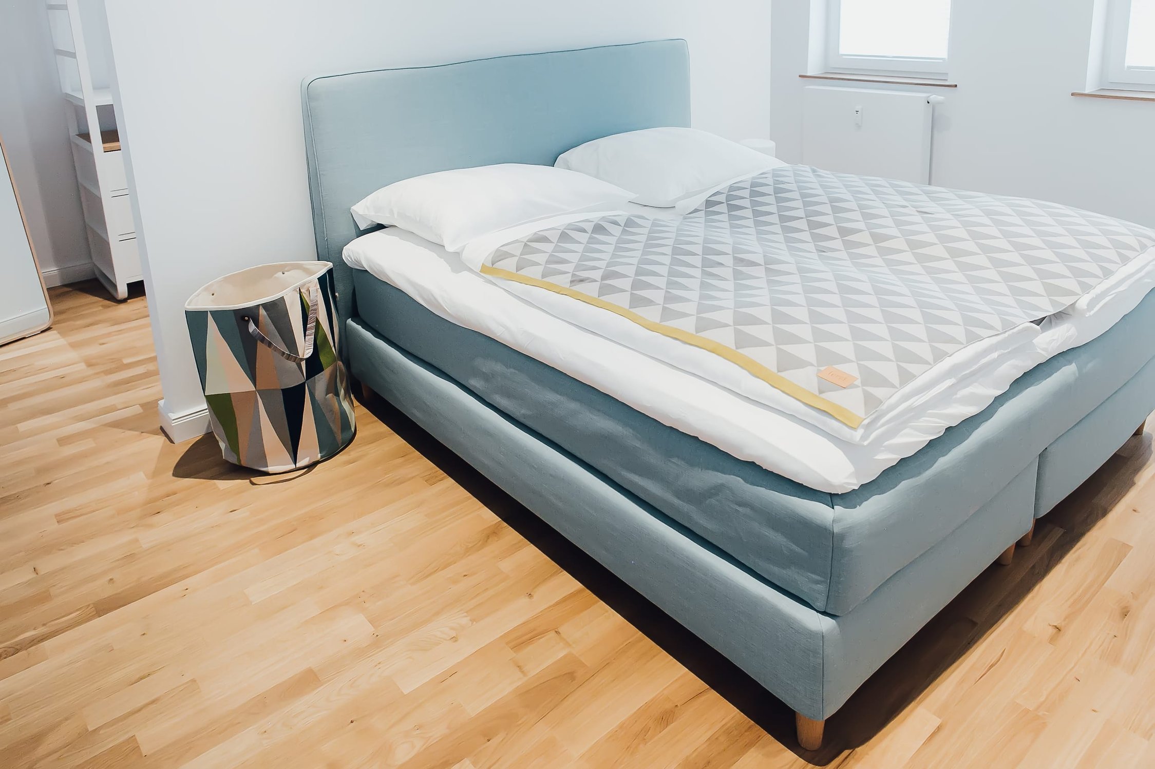 Schlafzimmer mit hellblauem Boxspringbett, hellem Holzboden und Nachttisch mit geometrischem Muster
