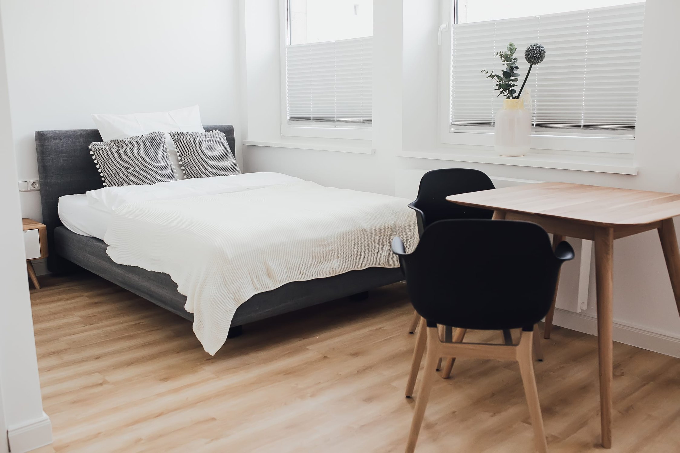 Schlafzimmer mit grauem Bett, weißer Bettwäsche, hellem Holzboden und Holztisch vor dem Fenster