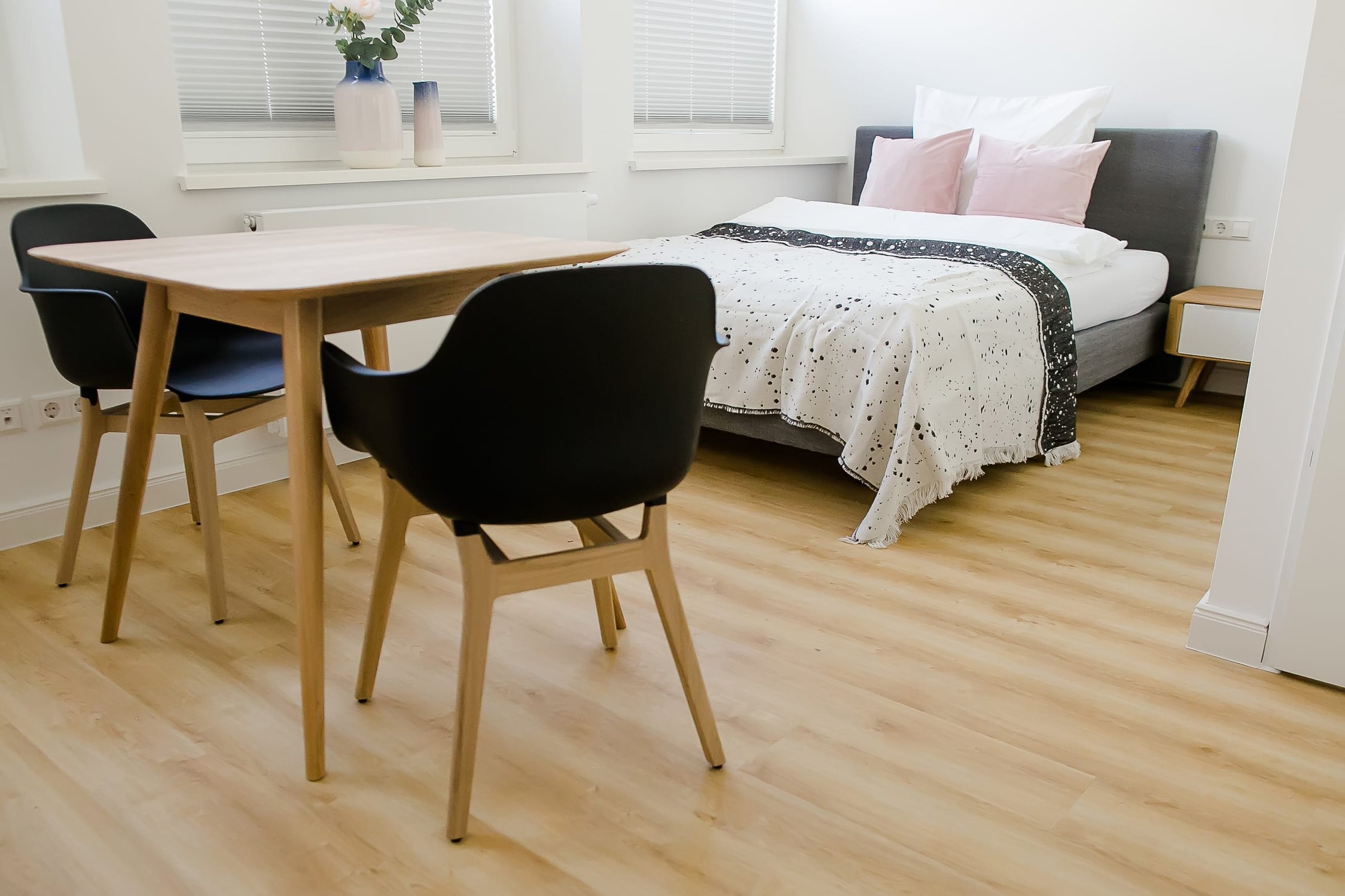 Heller Raum mit Bett, Holztisch, schwarzen Stühlen, zwei Fenstern und Holzfußboden