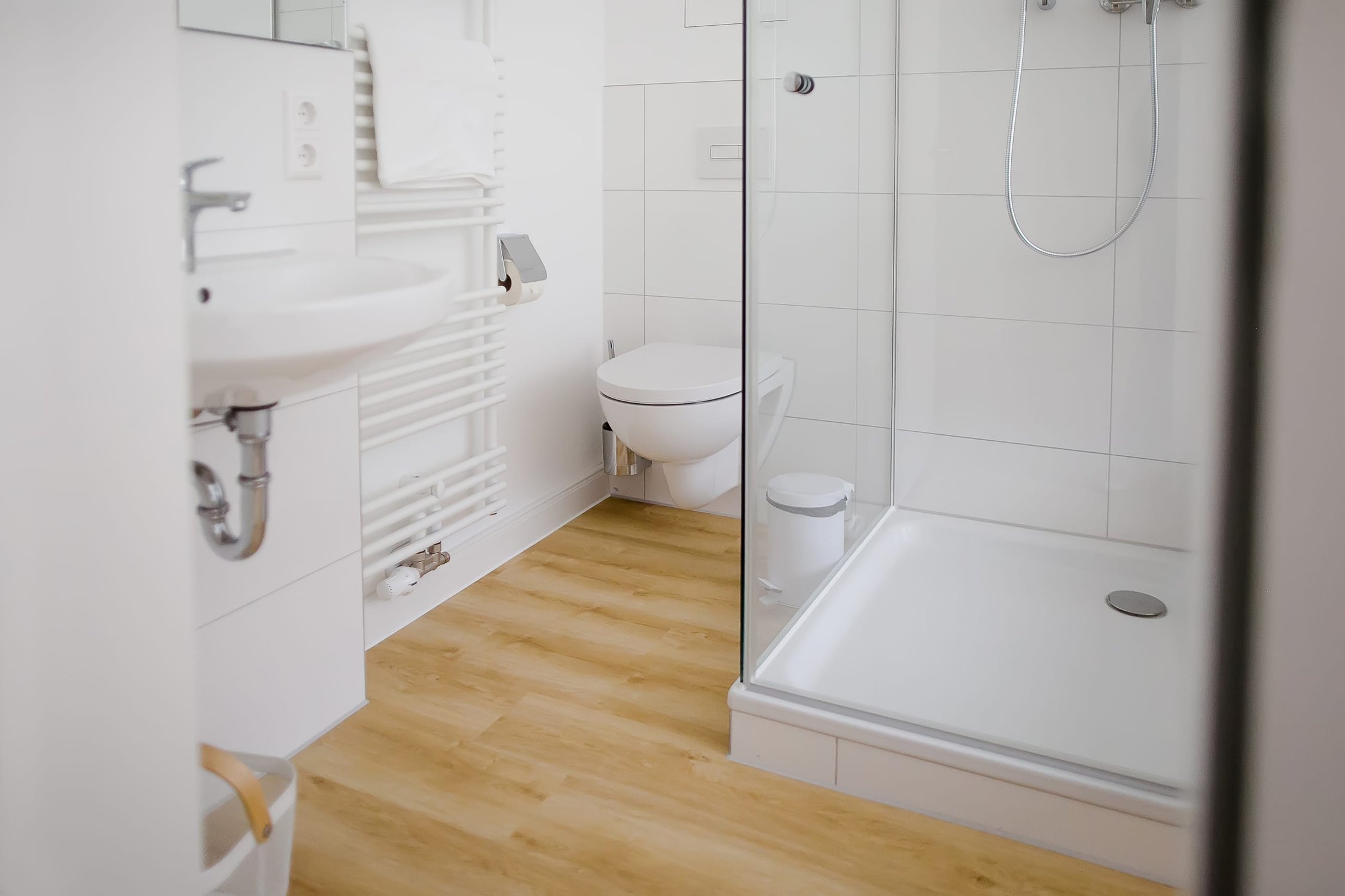 Helles Badezimmer mit weißen Fliesen, holzfarbenem Boden, Dusche und Hänge-WC