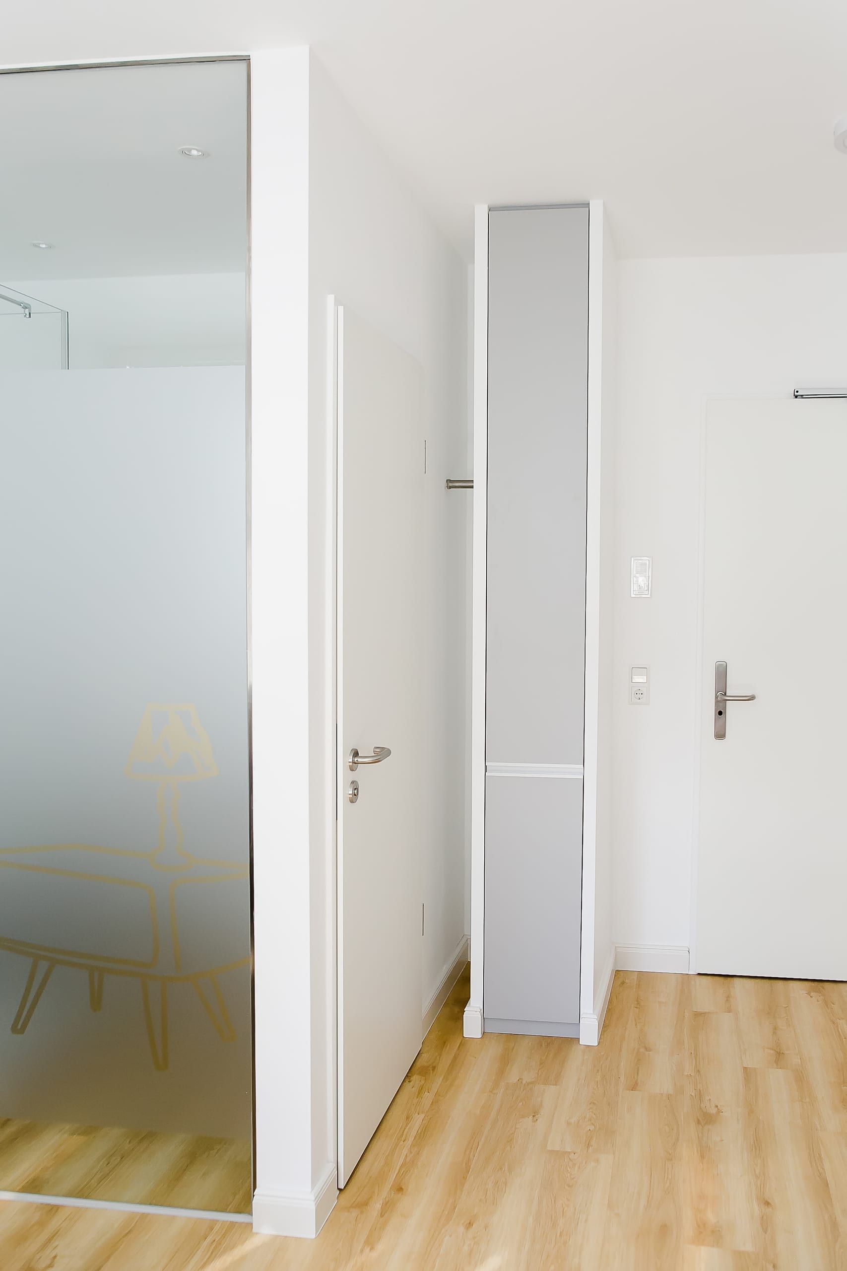 Heller Flur mit Holzboden, Milchglastür und weiß-grauem schmalen Schrank