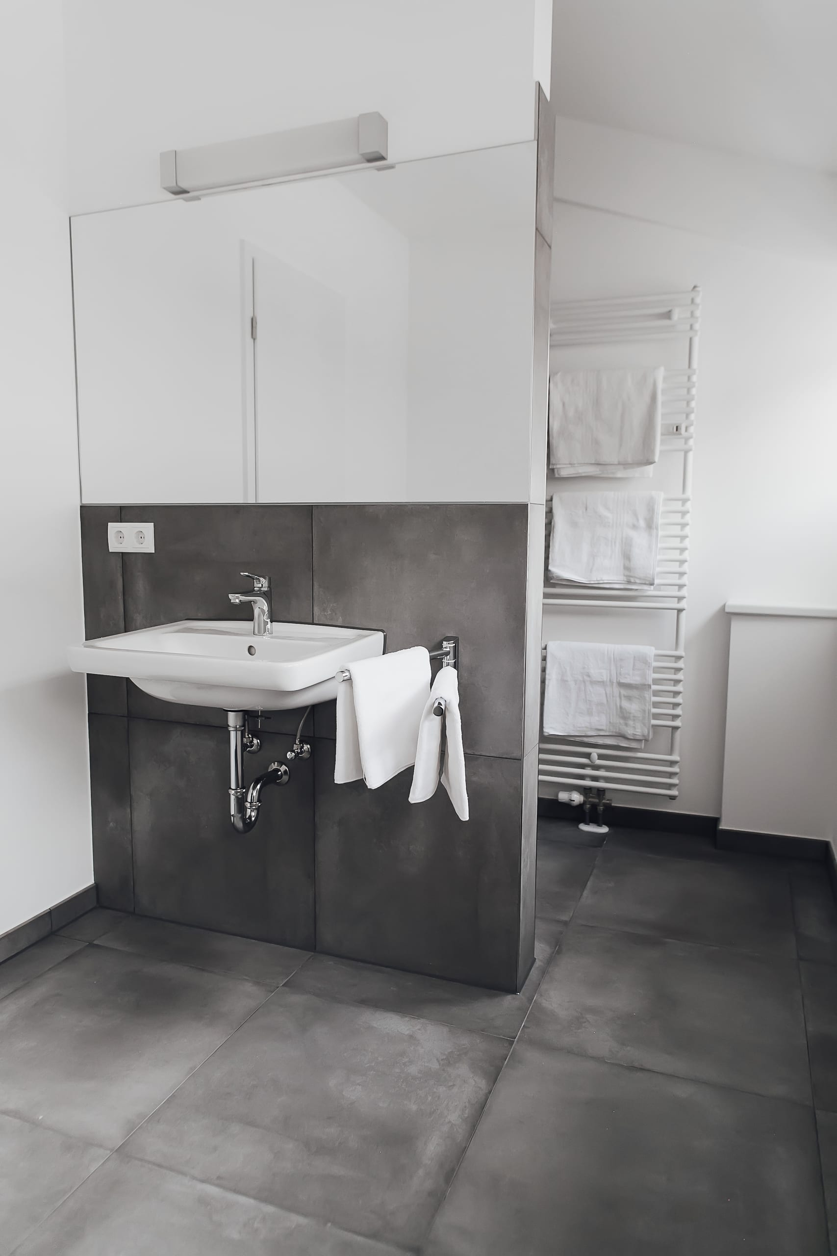 Badezimmer mit weißem Waschbecken, grauen Fliesen, Spiegel und Handtuchheizkörper