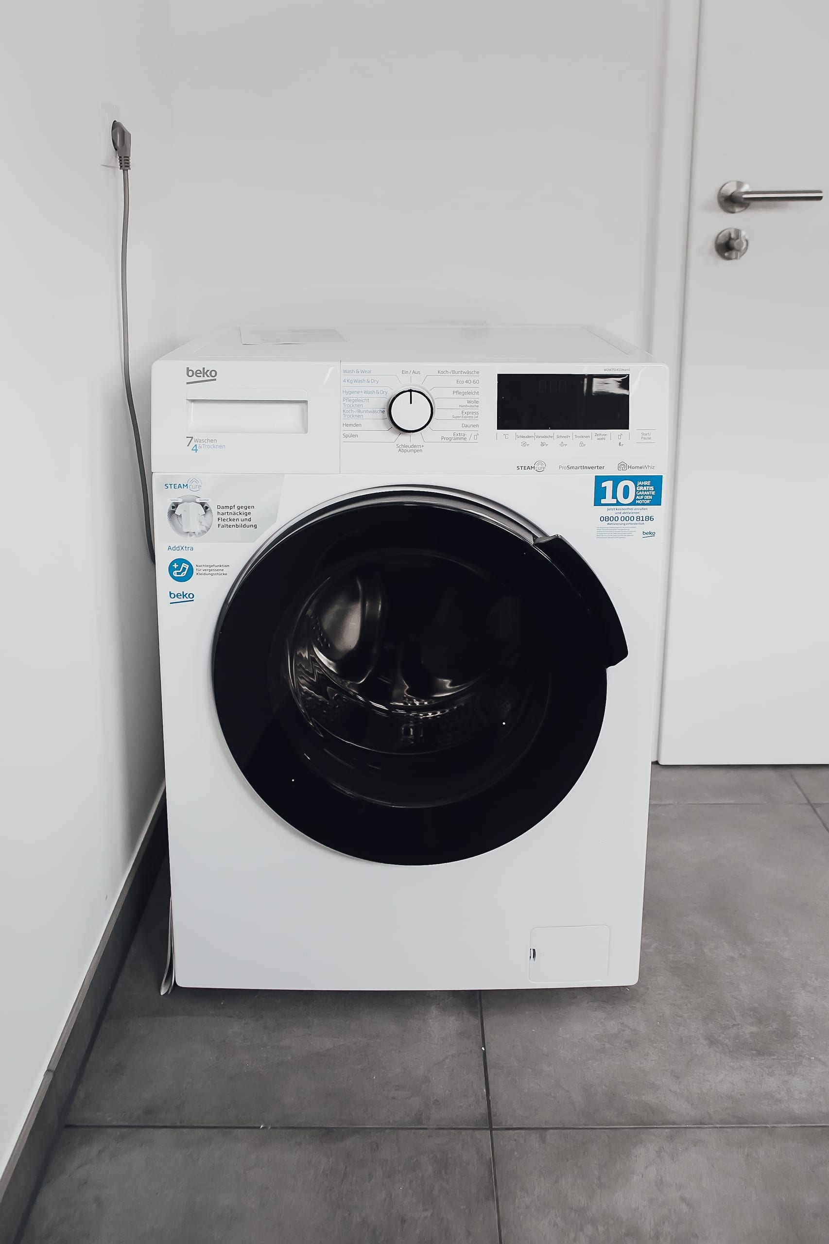 Weiße Waschmaschine mit schwarzer Trommelöffnung, vor weißer Wand und auf grauen Fliesen