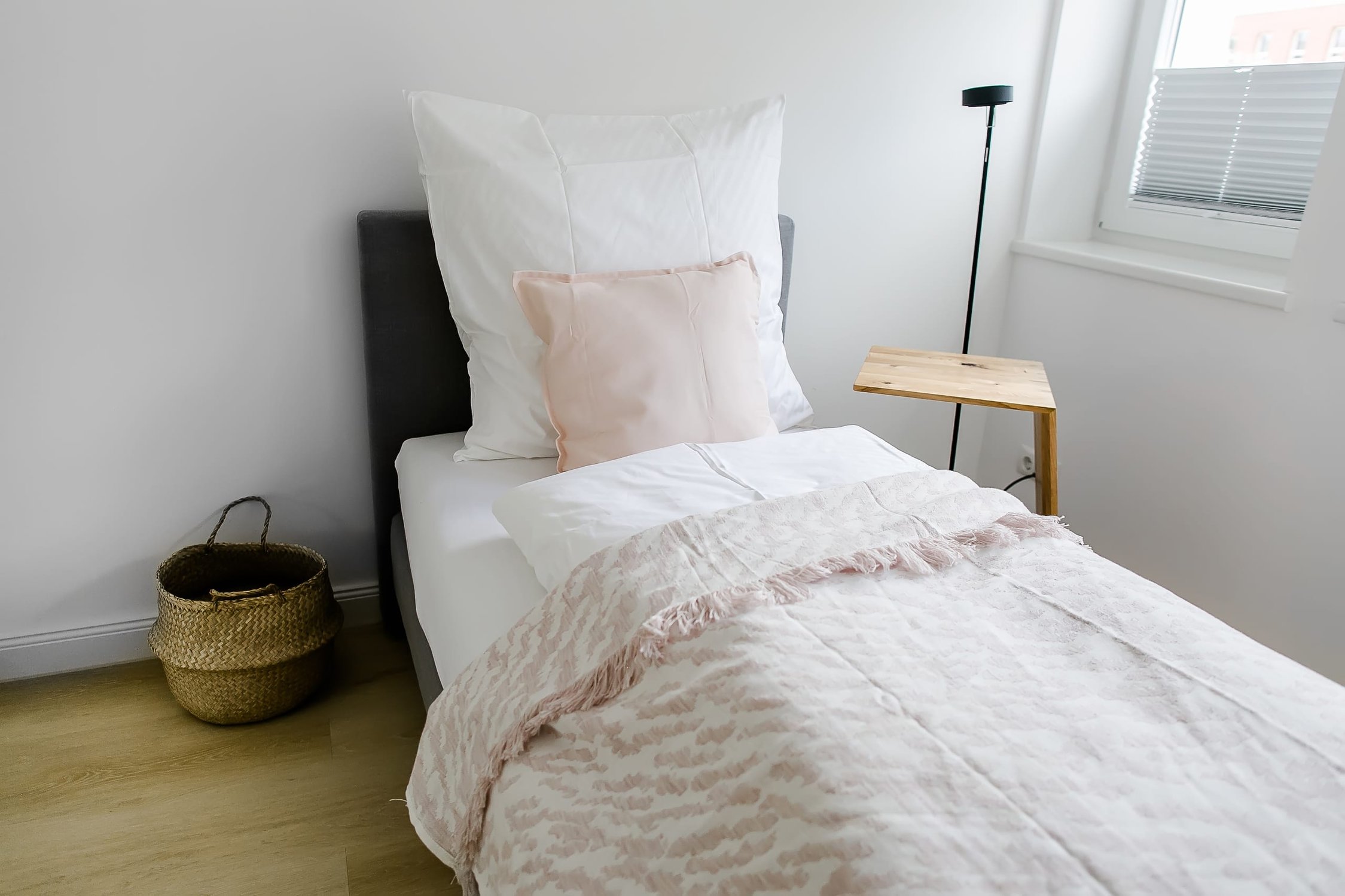 Helles Schlafzimmer mit Einzelbett, weißer Bettwäsche, rosa Kissen, Holztisch, Fenster
