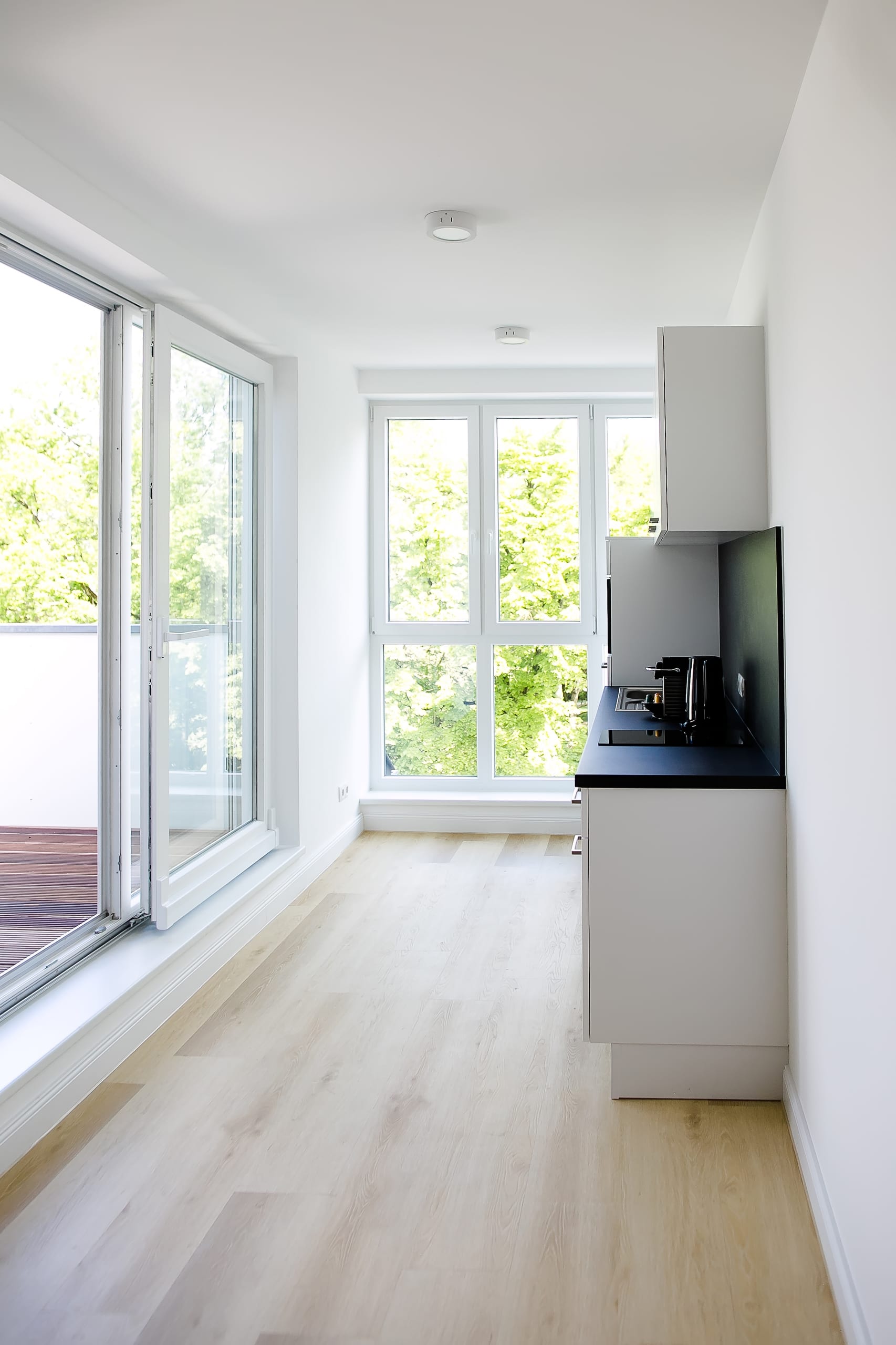 Schmaler Raum mit heller Holzoptik-Boden, weiße Küchenzeile, zwei großen Fenstern