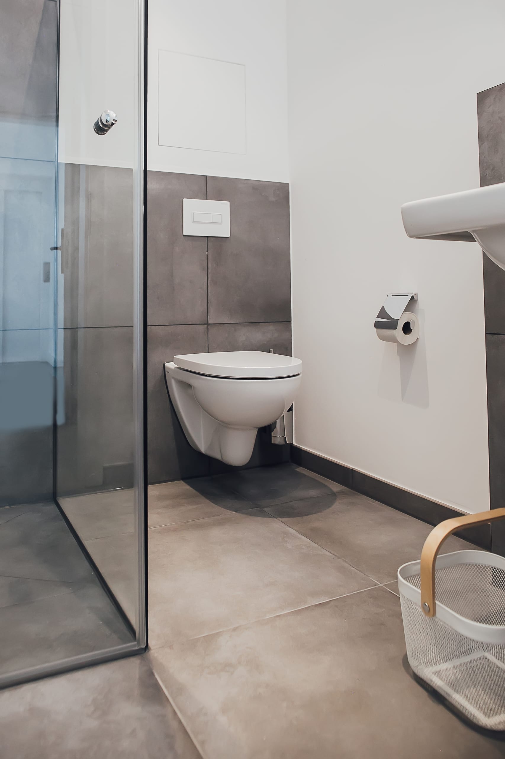 Badezimmer mit weißer Hänge-Toilette, grauen Fliesen, Glasduschwand und weißer Wäschekorb