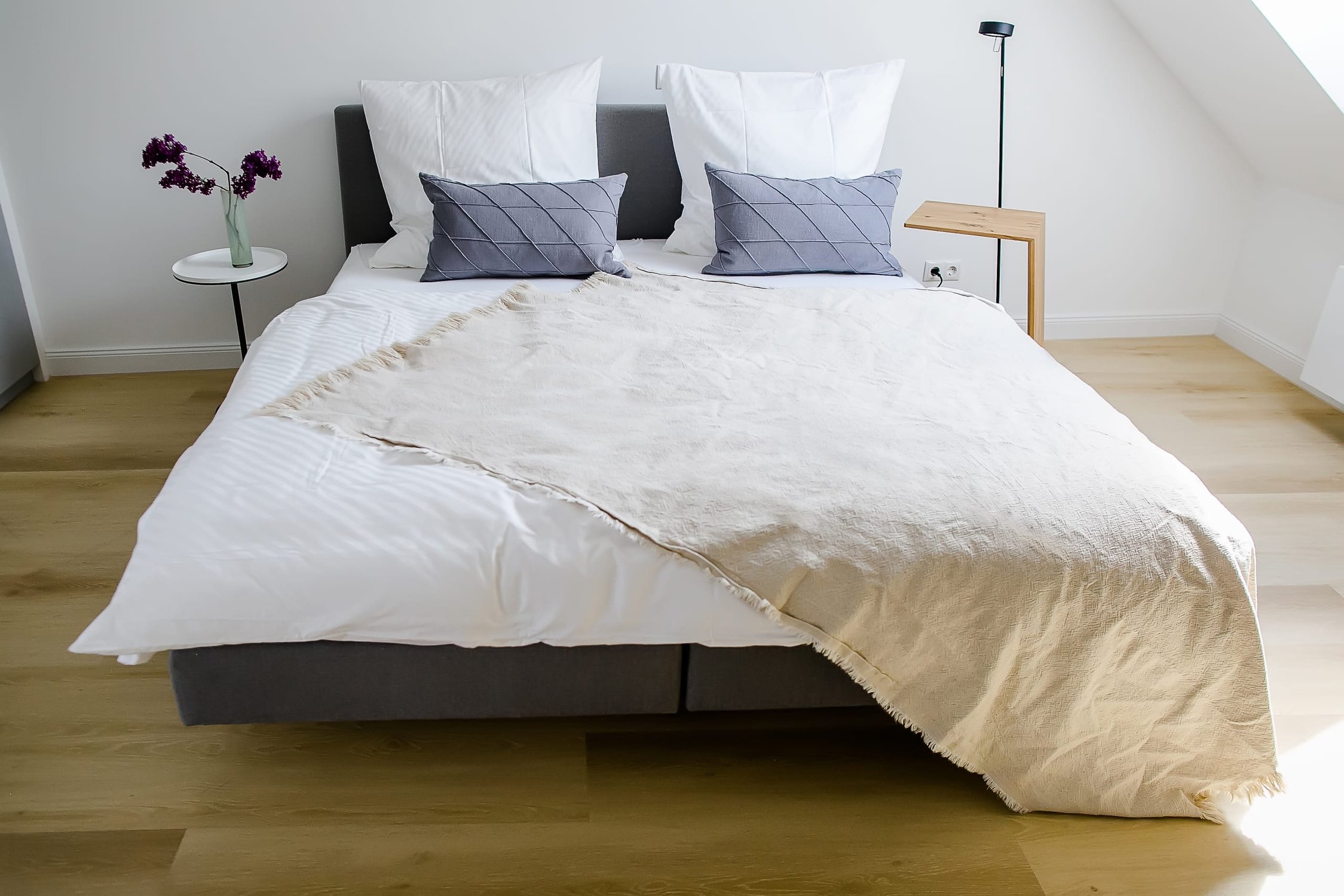 Helles Schlafzimmer mit Graubett, weißer Bettwäsche, beigem Plaid und zwei Nachttischen