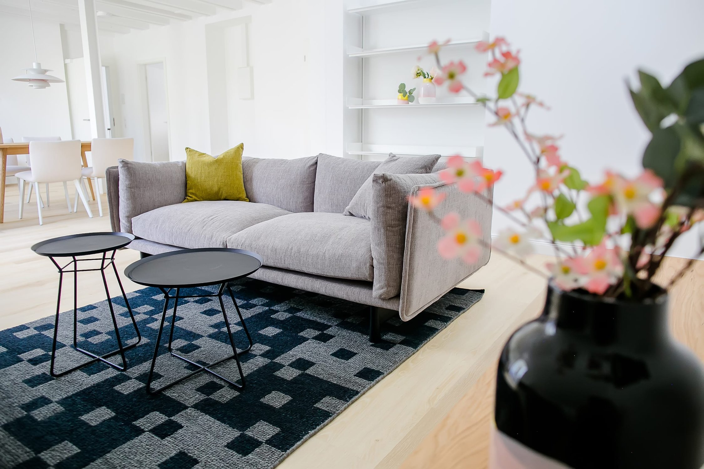Helles Wohzimmer mit grauem Sofa, schwarz-weißem Teppich, schwarzen Beistelltischen und Blumenvase