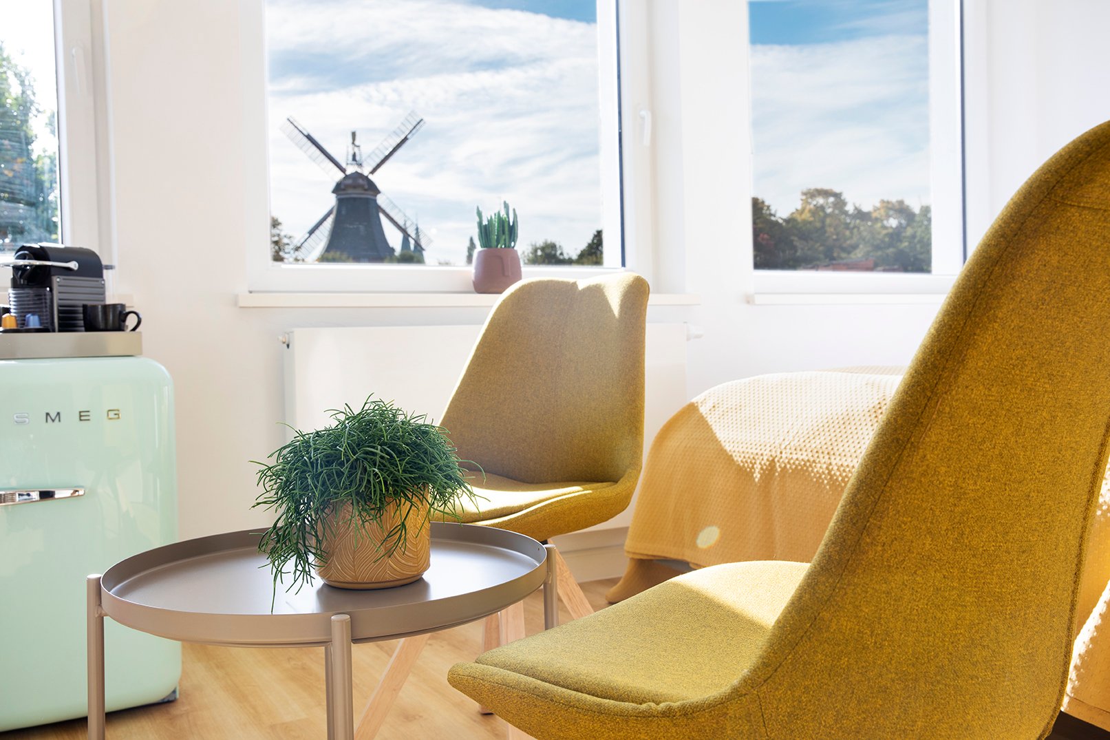 Moderner Wohnbereich mit mintgrünem Retro-Kühlschrank, zwei gelben Mid-Century-Stühlen, kleinem Beistelltisch mit grüner Pflanze und Blick auf eine Windmühle durch große Fenster