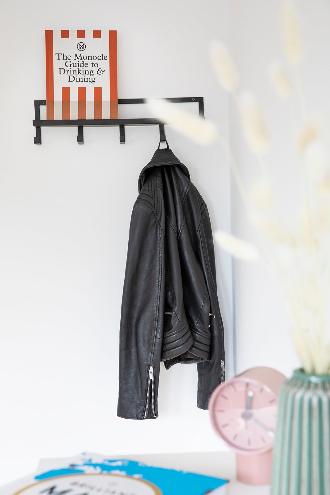 Schwarzes Wandregal mit Haken, an dem eine schwarze Lederjacke hängt, mit dem Buch 'The Monocle Guide to Drinking & Dining', unscharfe rosafarbene Uhr und Pflanze im Hintergrund