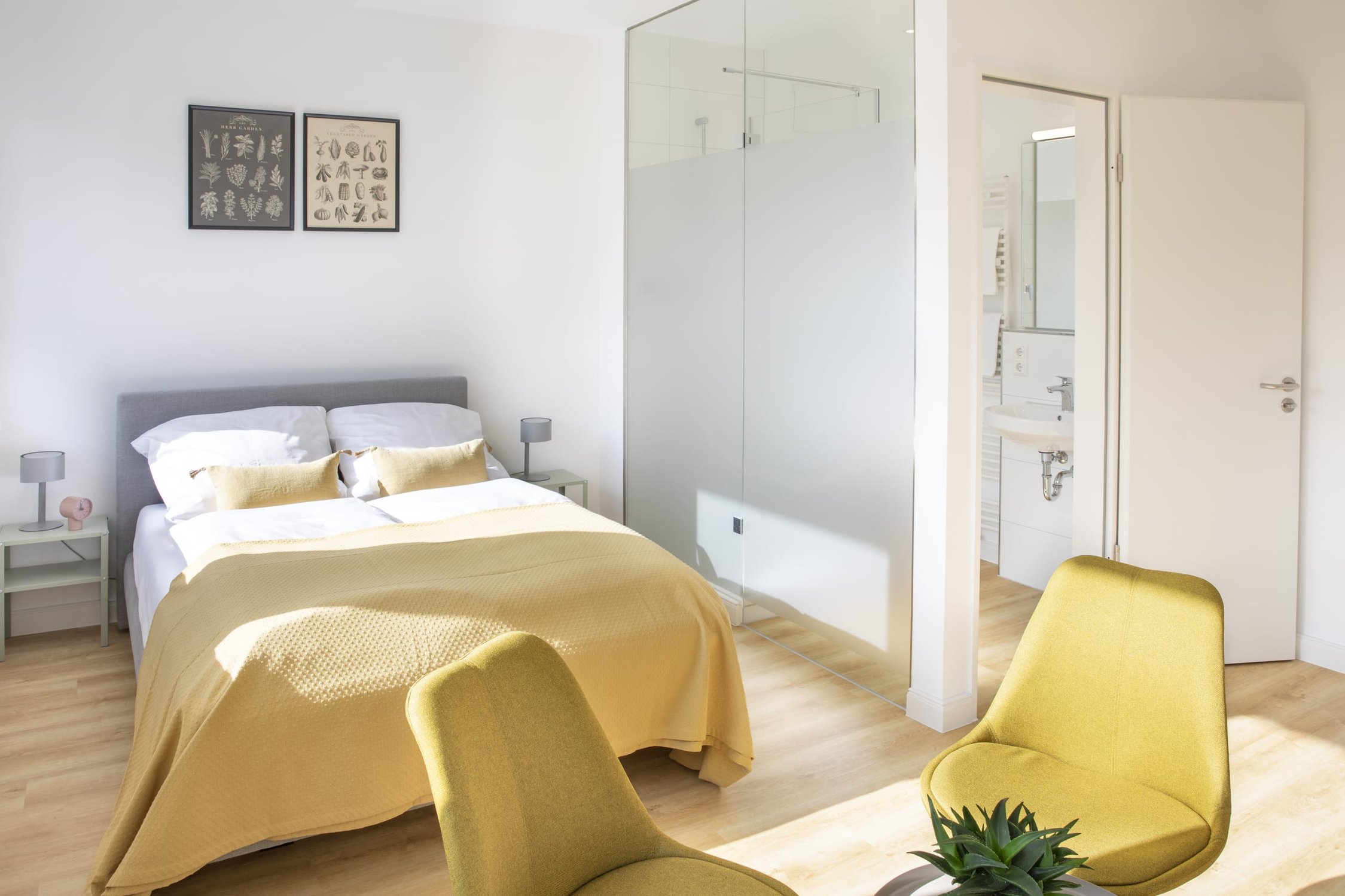 Helles Schlafzimmer mit Doppelbett, gelber Tagesdecke, zwei gelben Sesseln und Badezimmerzugang