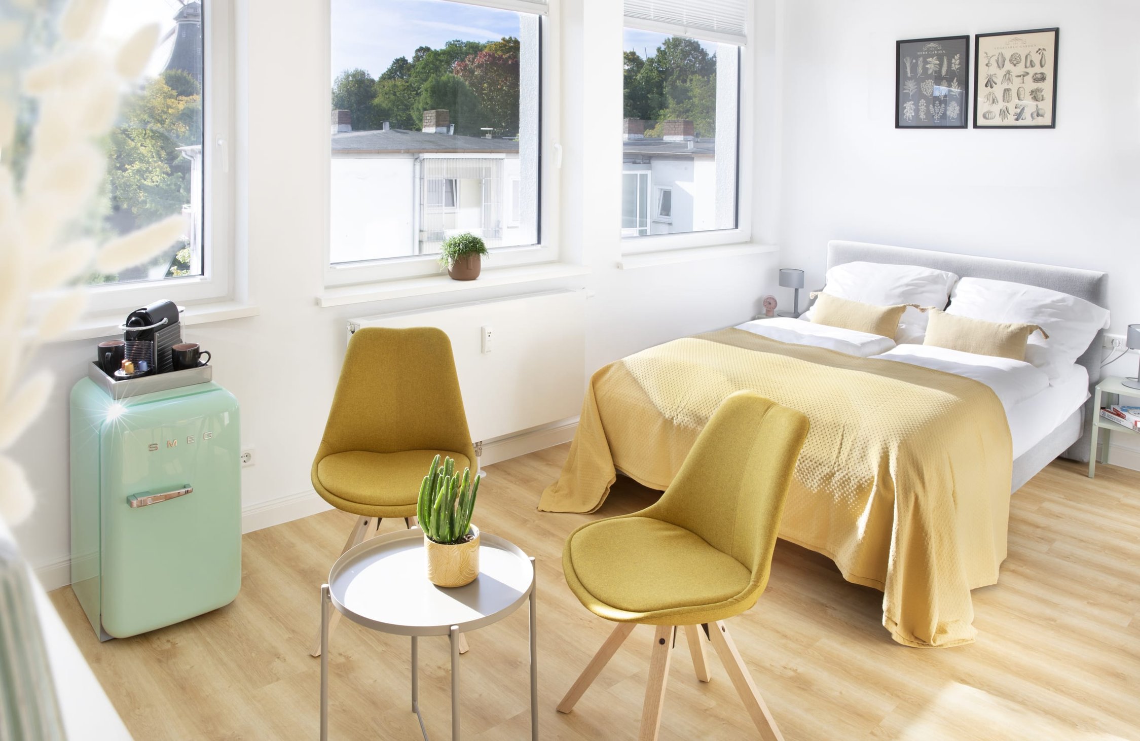 Helles Schlafzimmer mit mintgrünem Kühlschrank, zwei gelben Stühlen und Doppelbett mit gelber Decke