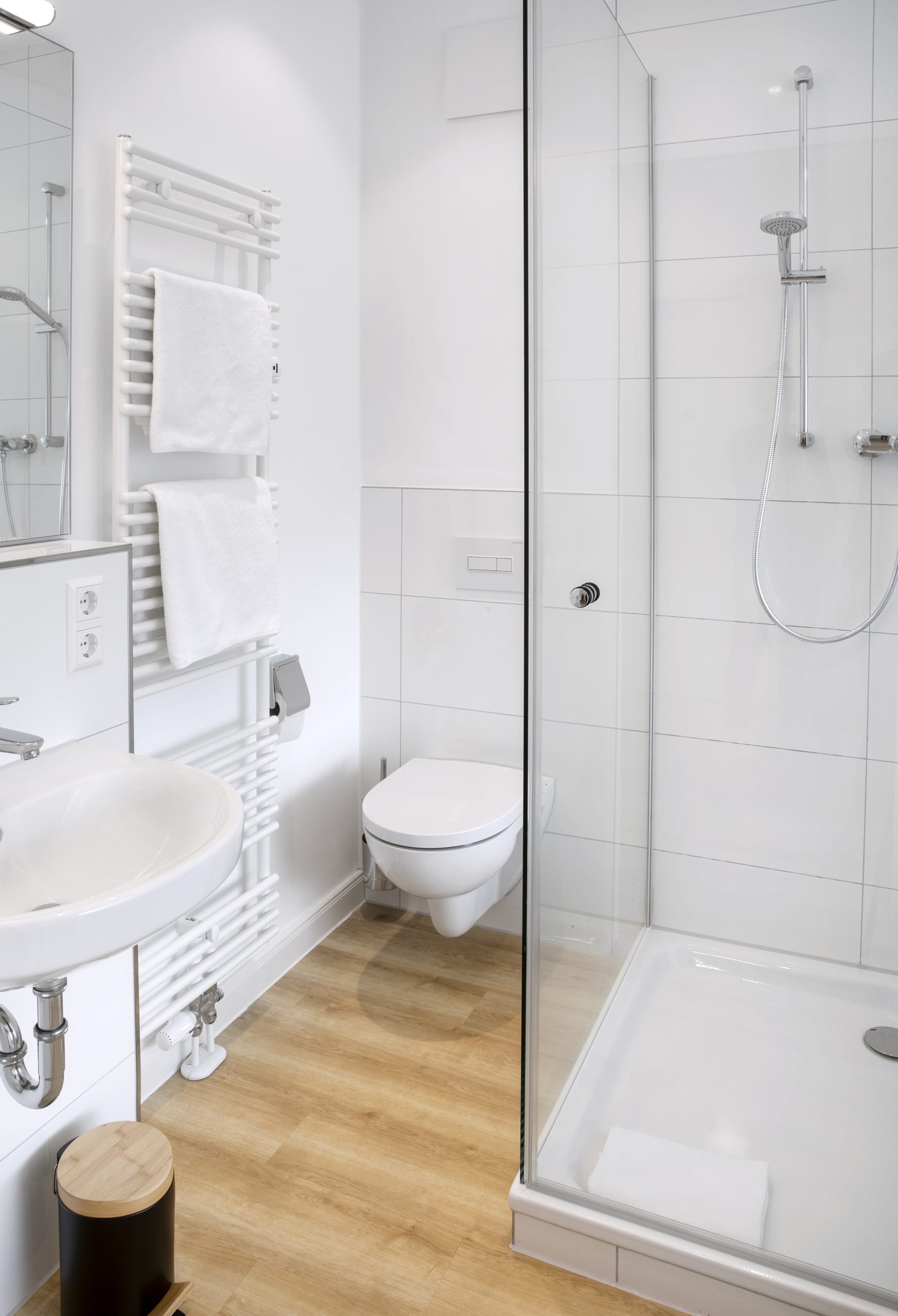 Helles Badezimmer mit weißen Fliesen, gläserner Dusche, weißer Toilette und Waschbecken, Holzfußboden