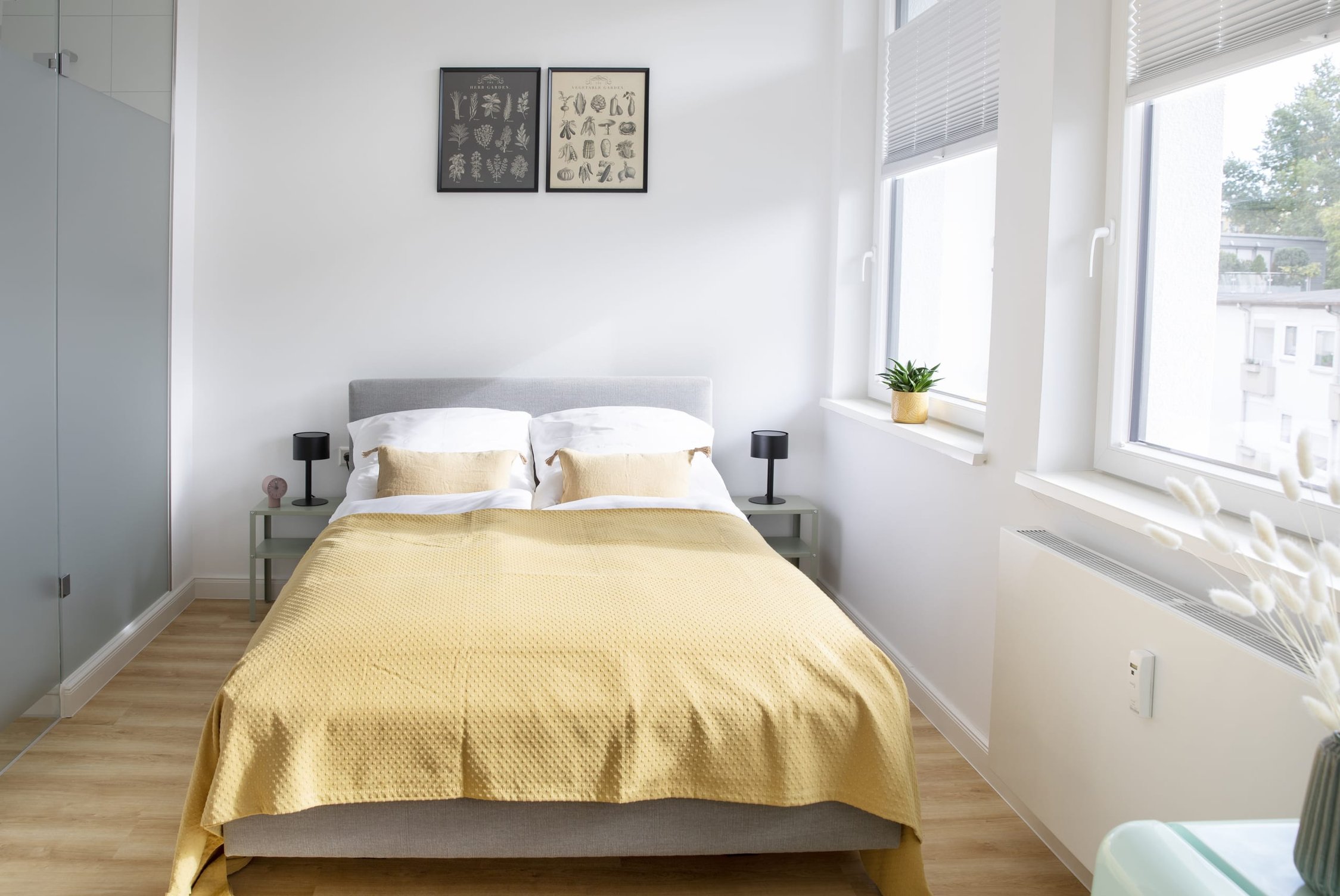 Helles Schlafzimmer mit grauem Bett, gelber Decke, zwei Fenstern und Schrank