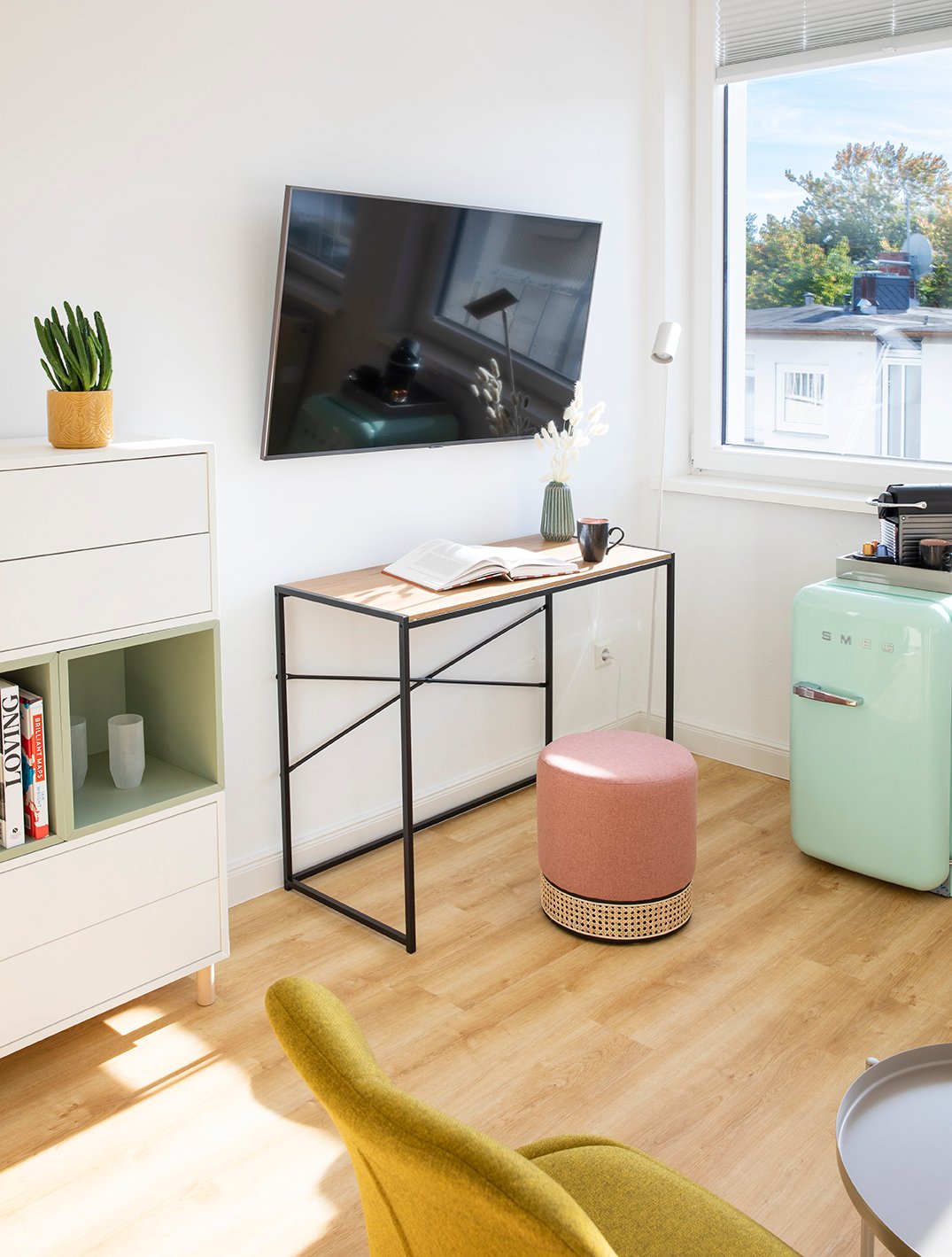 Modernes Schlaf-/Wohnzimmer mit weißen Lagermöbeln und Regal, schwarzem Wandfernseher, Schreibtisch mit schwarzem Metallgestell, rosafarbenem Hocker, mintgrünem Retro-Kühlschrank und hellem Holzboden